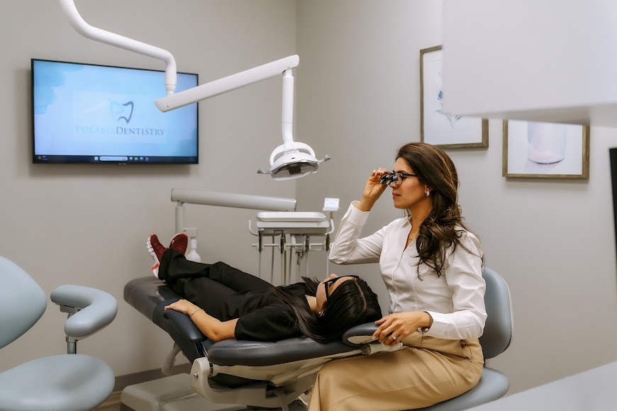 A dental professional beside a patient in a treatment chair, with an overhead light and wall-mounted TV